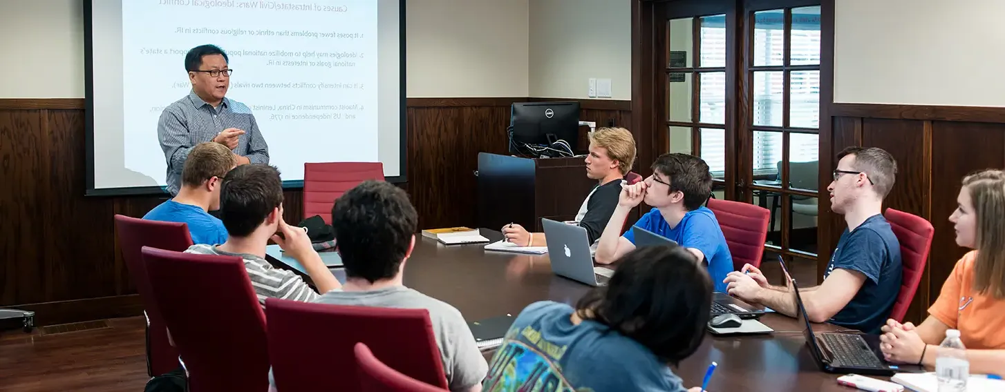 A group of students sitting around a boardroom style room during a presentation.